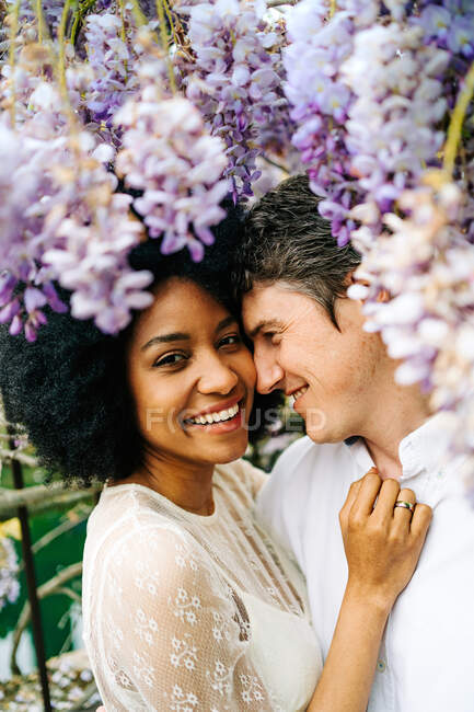 Vista lateral do casal multirracial amoroso abraçando no parque com flores de wisteria roxo florescendo no verão — Fotografia de Stock