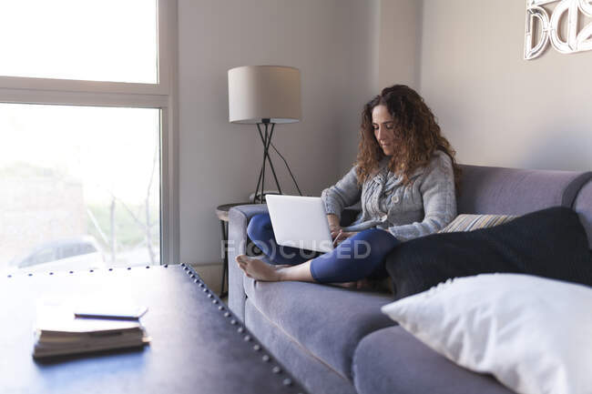 Seitenansicht einer Geschäftsfrau mit lockigem Haar, die auf dem Sofa sitzt und mit ihrem Laptop arbeitet — Stockfoto