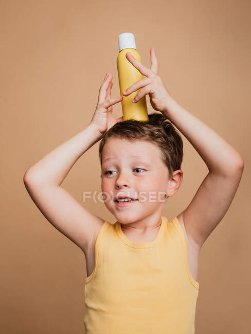 Cheerful preteen boy in swimsuit standing with bottle of sunblock cream on head on brown background in studio and looking away — Stock Photo
