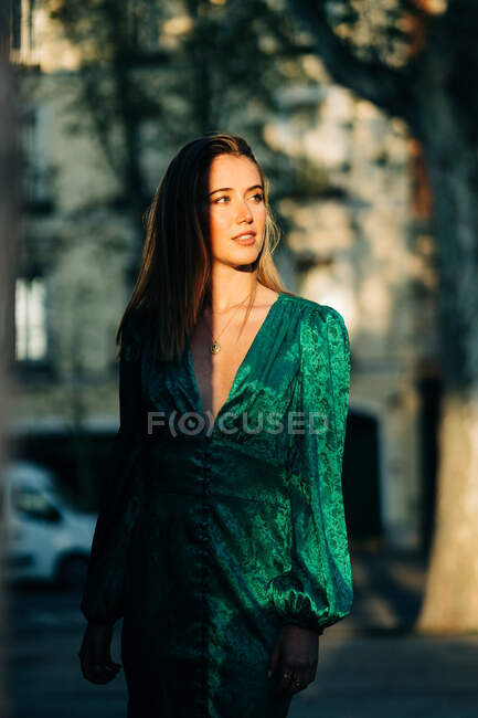 Carefree female in trendy green dress standing with one hand in her hair in street and looking away while a building shadows it — Stock Photo