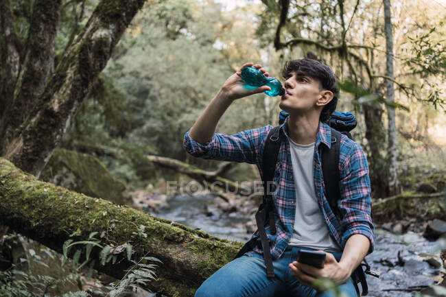 Durstige männliche Wanderer mit Rucksack trinken Wasser aus der Flasche, während sie auf einem Felsen in der Nähe des Wasserfalls im Wald sitzen und in der Pause wegschauen — Stockfoto