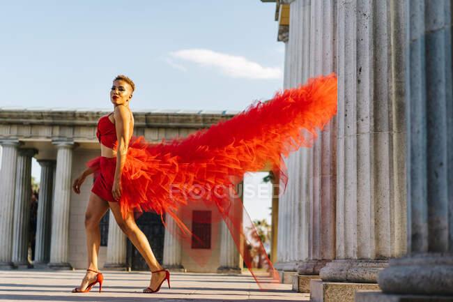 Вид сбоку на модную этническую женщину в красном платье с оборками и туфлями на высоком каблуке, прогуливающуюся по городским колоннам, глядя в камеру — стоковое фото