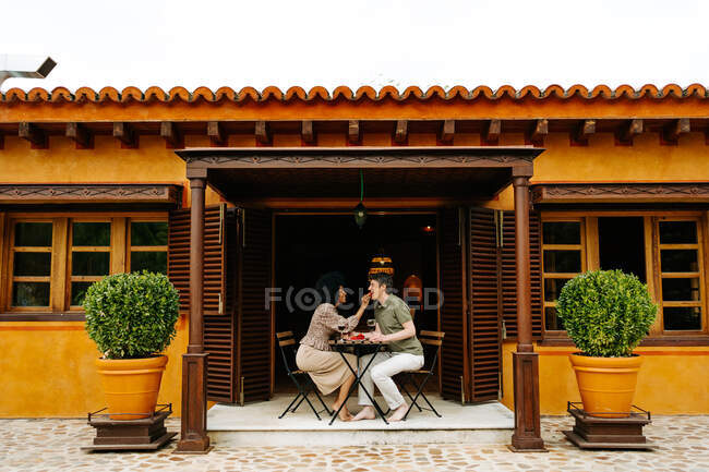 Side view of black woman feeding man with strawberry while sitting at table on terrace of cottage and enjoying romantic date at weekend — Stock Photo