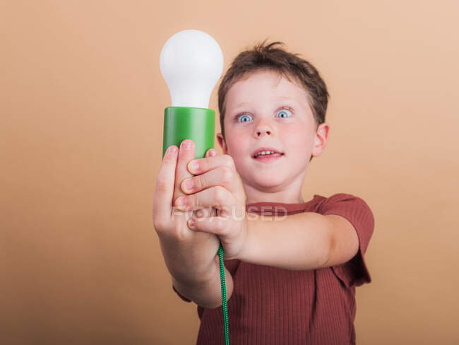 Удивленный ребенок в футболке с пластиковой лампочкой представляет идею концепции на бежевом фоне — стоковое фото