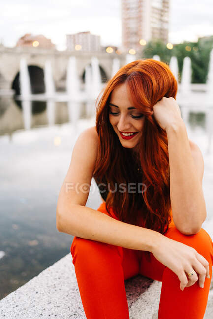 Cheerful female with long ginger hair and in bright orange pants sitting on border on promenade in city and leaning on hand — Stock Photo