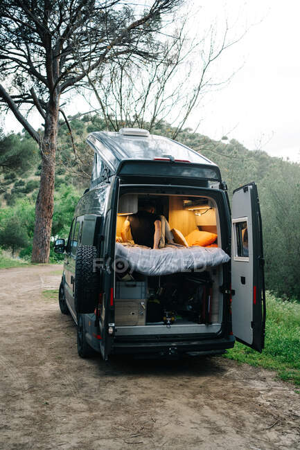 Pareja ambulante irreconocible sentada dentro de una moderna caravana estacionada en la verde zona montañosa en el campo de verano - foto de stock