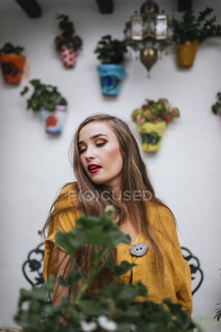 Mulher encantadora com cabelos longos e lábios vermelhos em pé no pátio com plantas verdes no verão e olhando para longe — Fotografia de Stock