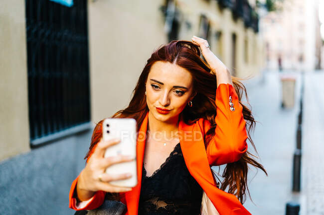 Mulher elegante com cabelo vermelho e em jaqueta laranja vibrante tomando auto tiro no smartphone na rua da cidade — Fotografia de Stock