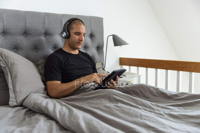 Hombre sereno en auriculares sentados en la cama debajo de la manta y navegando por las redes sociales en la tableta mientras escucha música por la mañana - foto de stock