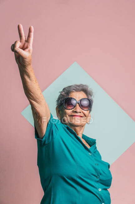 Весела сучасна літня жінка в модних сонцезахисних окулярах, що показують v знак і дивляться на камеру на двокольоровому фоні в студії — стокове фото