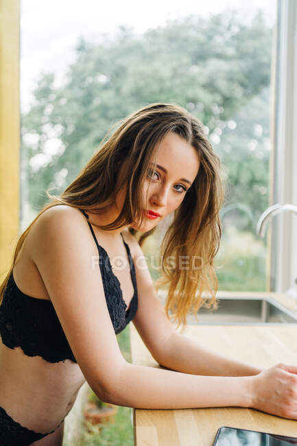 Vista lateral da fêmea sedutora em lingerie preta e com lábios vermelhos sentados no balcão na cozinha em casa e olhando para a câmera — Fotografia de Stock