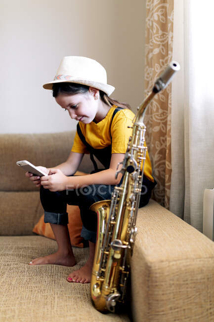 Kind mit Hut SMS auf Handy, während es mit Saxophon auf Couch im Wohnzimmer sitzt — Stockfoto