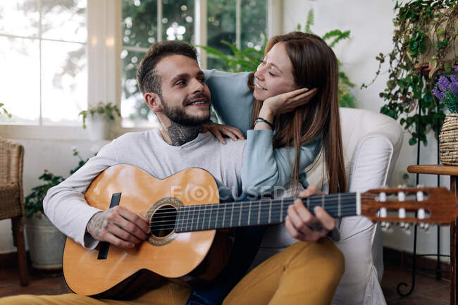 Fröhlich tätowierte männliche Musiker spielen Gitarre in der Nähe zufriedener weiblicher Geliebter, während sie einander im Sessel im Hauszimmer anschauen — Stockfoto