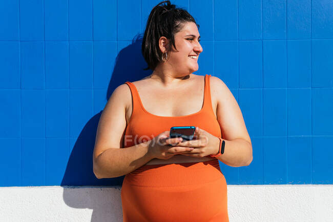 Fröhliche Plus-Size-Ethnie-Athletin in aktiver Kleidung mit Handy, das im Sonnenlicht vor blau gekachelter Wand wegschaut — Stockfoto