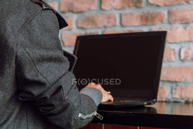 Rückseite Ernte Frau mit gefärbten Haaren in stilvollem Mantel sitzt am Tisch in der Nähe Ziegelwand, während mit Laptop — Stockfoto
