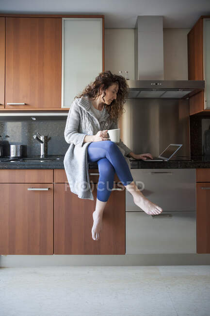 Ділова жінка з кучерявим волоссям сидить на кухні, приймаючи настій, використовуючи свій ноутбук і працюючи вдома — стокове фото