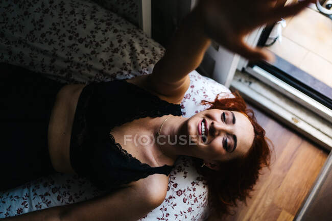 Вид сверху жизнерадостной рыжеволосой женщины, лежащей на мягкой кровати с закрытыми глазами и протягивающей руку к камере дома — стоковое фото