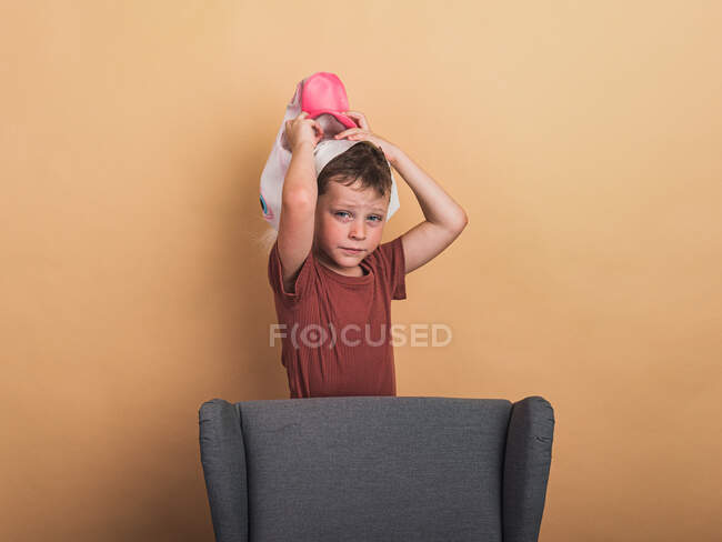 Exhausted child in t shirt taking off decorative mask from head while looking at camera on beige background — Stock Photo