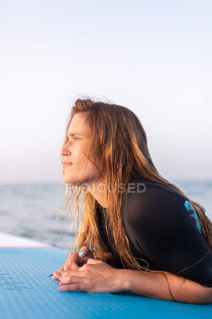 Vista lateral do surfista fêmea deitado na placa SUP e flutuando na água calma do mar no dia ensolarado olhando para longe — Fotografia de Stock