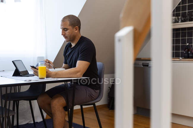 Взрослый мужчина просматривает мобильный телефон во время сидения за столом на кухне и завтрака утром дома и пить апельсиновый сок — стоковое фото