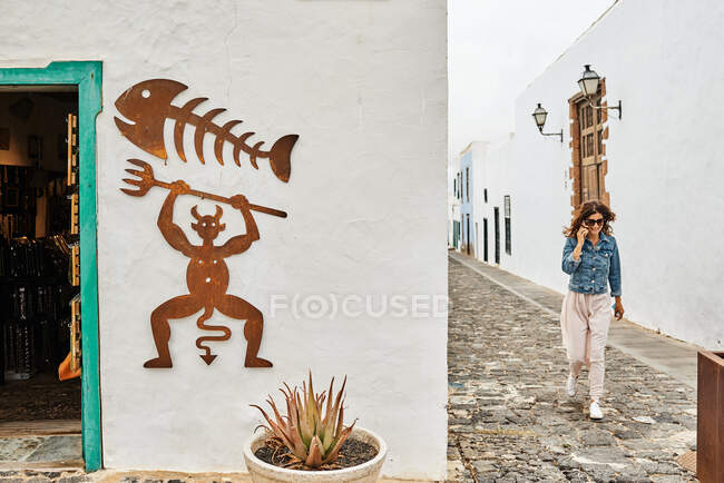 Frohe Frau in legerer Kleidung, die Telefonanrufe entgegennimmt, während sie auf einem Steinweg in der Nähe von Gebäuden mit Teufels- und Fischskelettbildern auf der Stadtstraße in Fuerteventura, Spanien, spaziert — Stockfoto