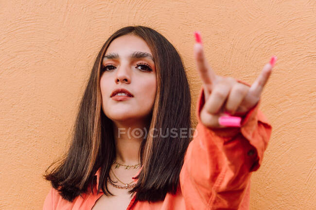 Charismatic confident female with long brown hair and makeup showing horn gesture against painted orange background — Stock Photo