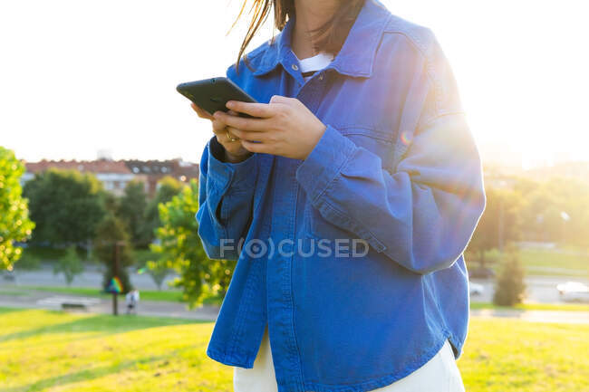 Gestutzte, bis zur Unkenntlichkeit gestylte Frau steht auf grünem Grashügel und surft an sonnigen Tagen auf dem Smartphone — Stockfoto