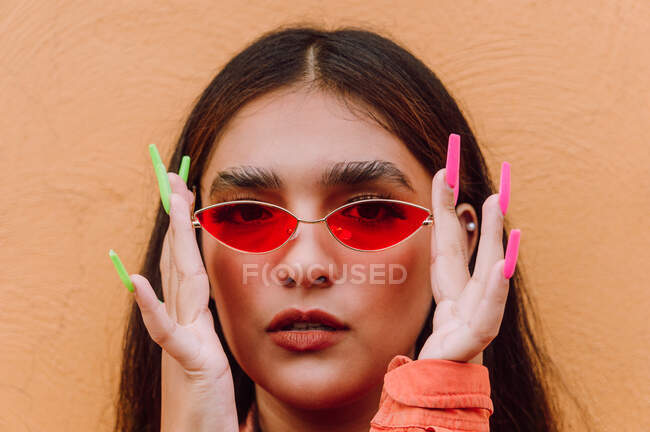 Retrato de corte de hembra carismática con uñas largas y brillantes que se ponen gafas de sol de moda contra la pared naranja mirando a la cámara - foto de stock