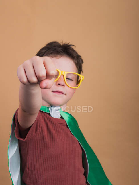 Ребенок в плаще супергероя и декоративных очках, показывающих жесты силы, глядя в камеру — стоковое фото
