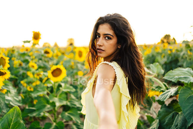 На вигляд молодої іспаномовної жінки в жовтому вбранні, яка дивиться на камеру і простягає руку, як запрошує слідувати за нею в безконечному полі з квітучими соняшниками в літній день. — стокове фото