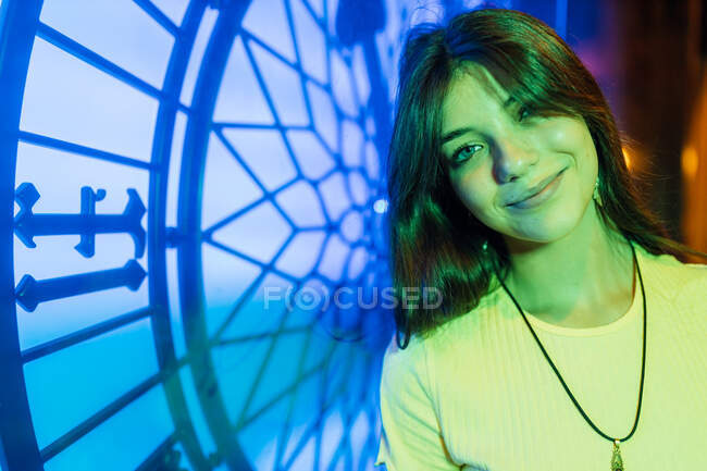 Young gentle female in pendant and earring with long hair looking at camera while she is illuminated with green neon lights in evening — Stock Photo