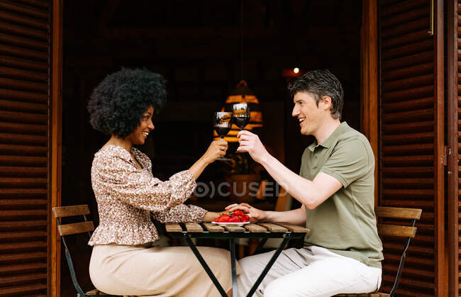Vista lateral do alegre casal multirracial de mãos dadas e copos de vinho tinto enquanto propõe brinde e desfruta de data romântica no terraço da casa à mesa com prato de morangos — Fotografia de Stock