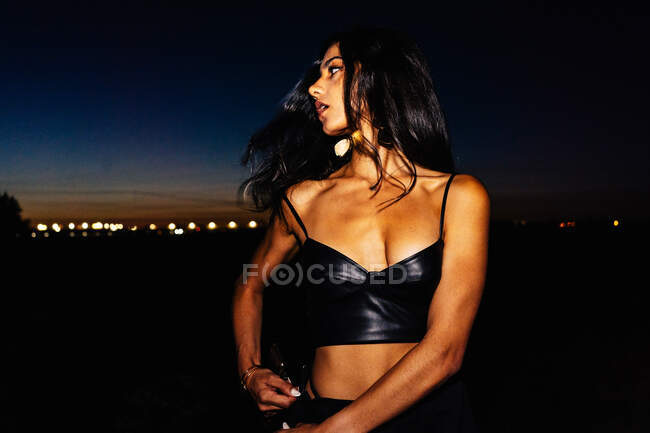 Long haired brunette in black leather top and sexy lingerie standing provocatively in dark field with glowing lights in twilight time — Stock Photo