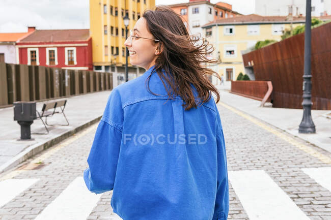 Back view of smiling female with flying hair walking along road in city on windy day — Stock Photo