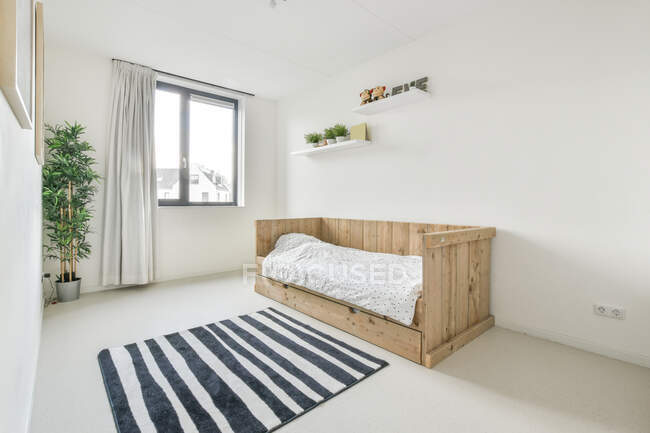 Интерьер современной белой спальни минималистского стиля с экологически чистым дизайном, включая односпальную деревянную кровать и полосатый ковер с растениями в горшке — стоковое фото