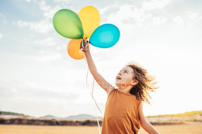Criança étnica sorridente com cabelo encaracolado brincando com balões de ar coloridos no campo de verão e olhando para cima — Fotografia de Stock