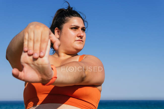 Atenta atleta étnica femenina con cuerpo curvilíneo haciendo ejercicio con los brazos extendidos mientras mira hacia otro lado contra el mar bajo el cielo azul - foto de stock