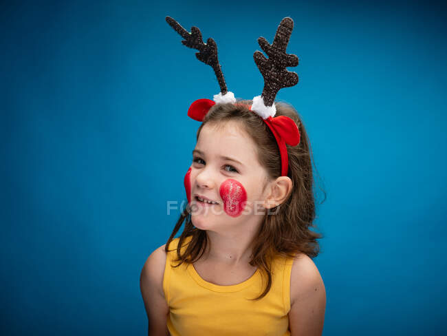 Smiling girl with cheeks painted red wearing toy deer horns and ears and looking at camera on blue background — Stock Photo