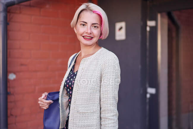 Альтернативная женщина с короткими окрашенными волосами, смотрящая в камеру и улыбаясь на городской улице против здания — стоковое фото