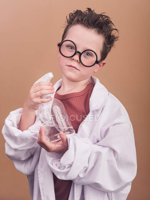 Enfant chimiste en lunettes décoratives et peignoir de laboratoire regardant la caméra avec de l'eau dans un flacon distributeur sur fond beige — Photo de stock