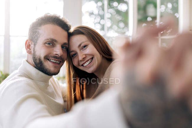 Веселый бородатый мужчина с искренней возлюбленной, делающей автопортрет, глядя в камеру дома — стоковое фото