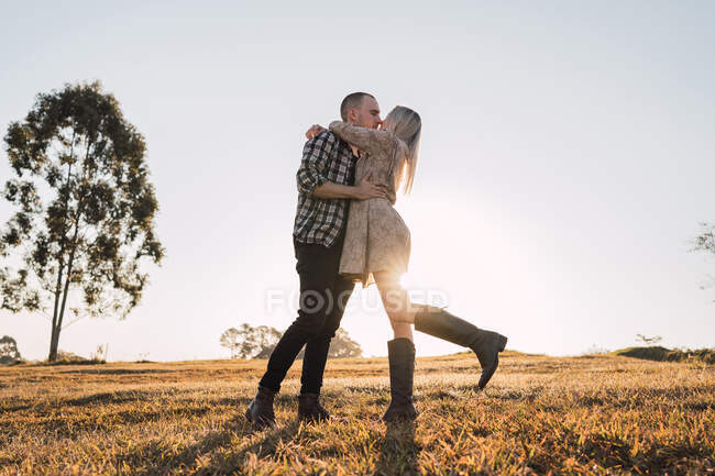 Low angle side view of loving couple embracing and kissing while standing on meadow on background of sunset sky — Stock Photo