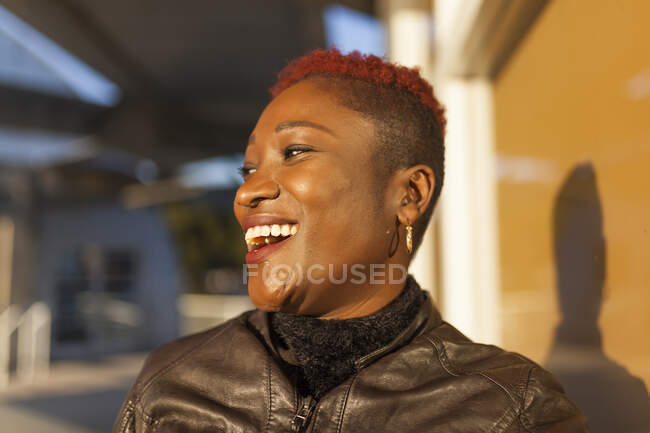 Vue latérale de la belle femme afro posant près de la fenêtre et souriant tout en regardant loin par une journée ensoleillée — Photo de stock