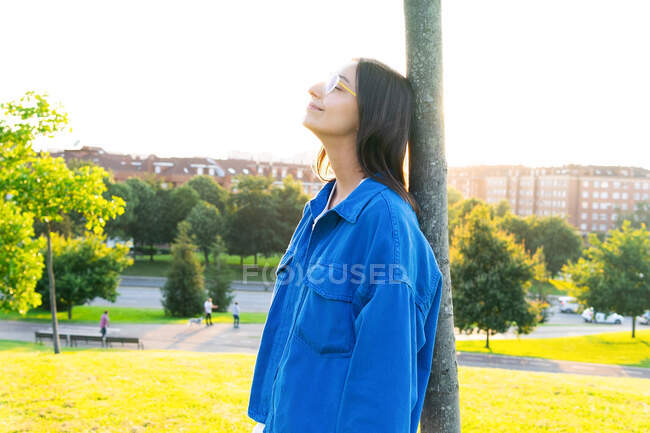 Seitenansicht einer friedlichen Frau in stylischem Outfit, die sich mit geschlossenen Augen auf einen Baumstamm lehnt, während sie im Gegenlicht auf dem grünen Rasen gegen die Stadt steht — Stockfoto
