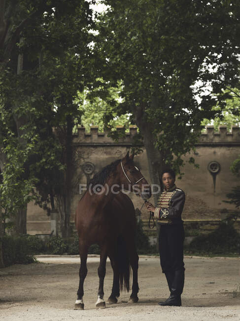 Volle Länge der erwachsenen schwarzen Hündin in elegantem Outfit, die mit braunem Pferd in der Nähe von grünen Bäumen und Burgzaun am Tag im Hof steht — Stockfoto