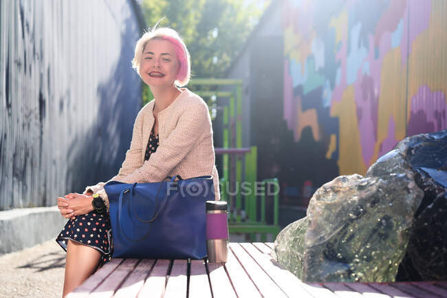Вид сбоку трехлетней альтернативной женщины с окрашенными волосами, сидящей на скамейке в городской зоне в городе — стоковое фото
