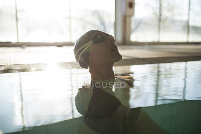 Jeune belle femme sur le bord de la piscine intérieure, avec maillot de bain noir, rayons de soleil entrant par la fenêtre — Photo de stock
