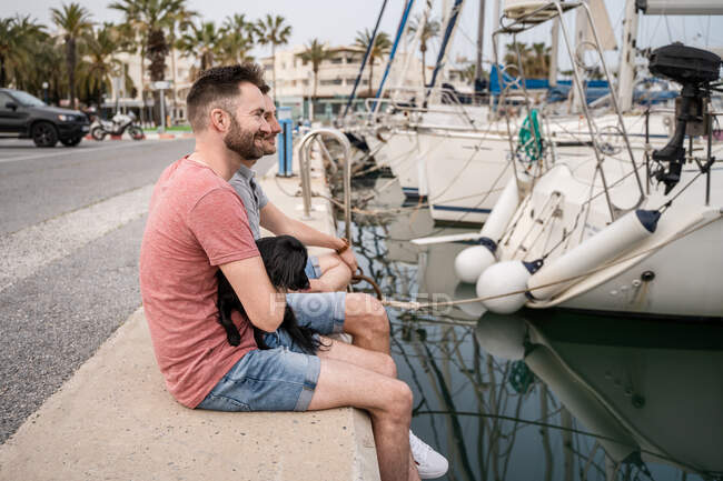 Боковой взгляд на собаку между веселым бородатым мужчиной, который обнимает анонимного гомосексуального партнера, разговаривая и сидя на пирсе в гавани — стоковое фото