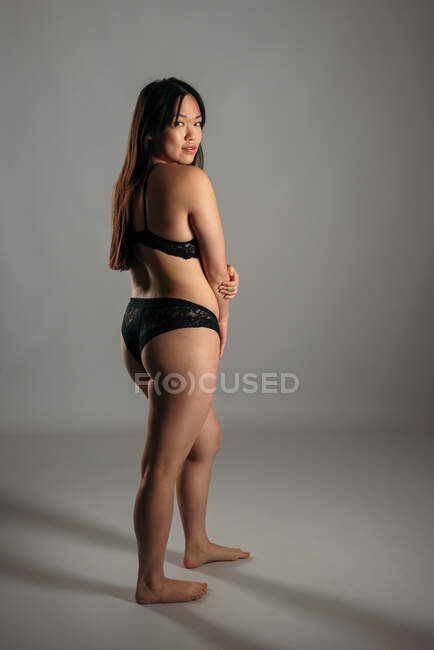 Mulher asiática confiante em lingerie preta em pé sobre fundo cinza em estúdio e olhando para a câmera — Fotografia de Stock