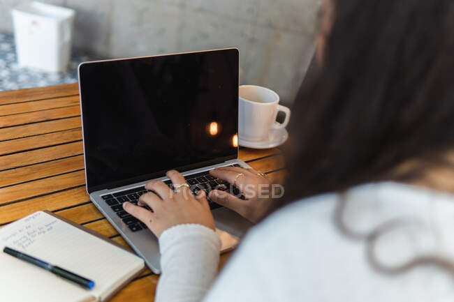 Обрезанная неузнаваемая молодая латиноамериканская студентка читает информацию на экране ноутбука при подготовке к университетскому экзамену в уютном кафе — стоковое фото